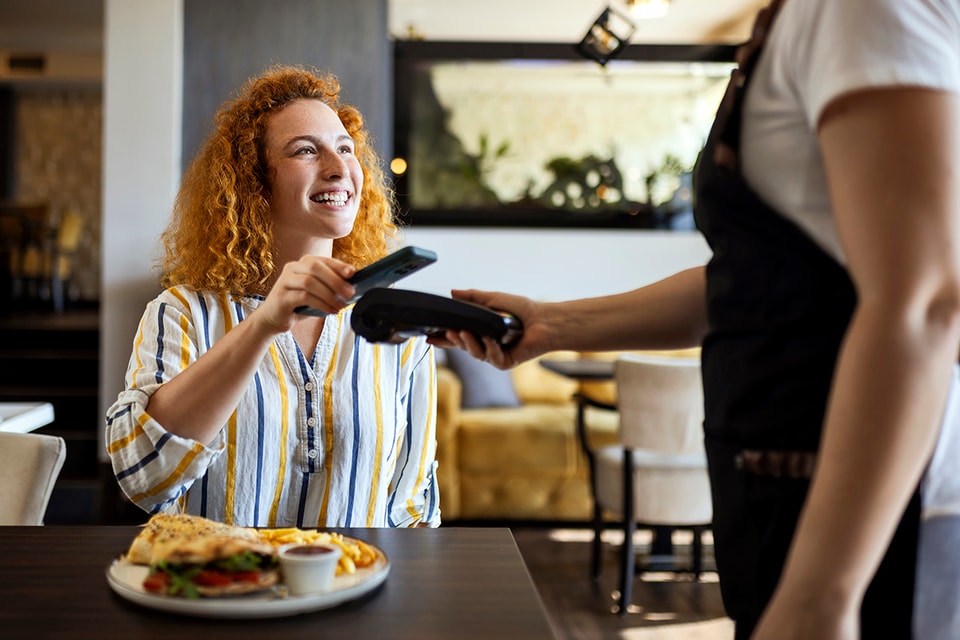 Dame betaler med mobiltelefonen på restaurant og smiler, til illustrasjon for Samsung Pay